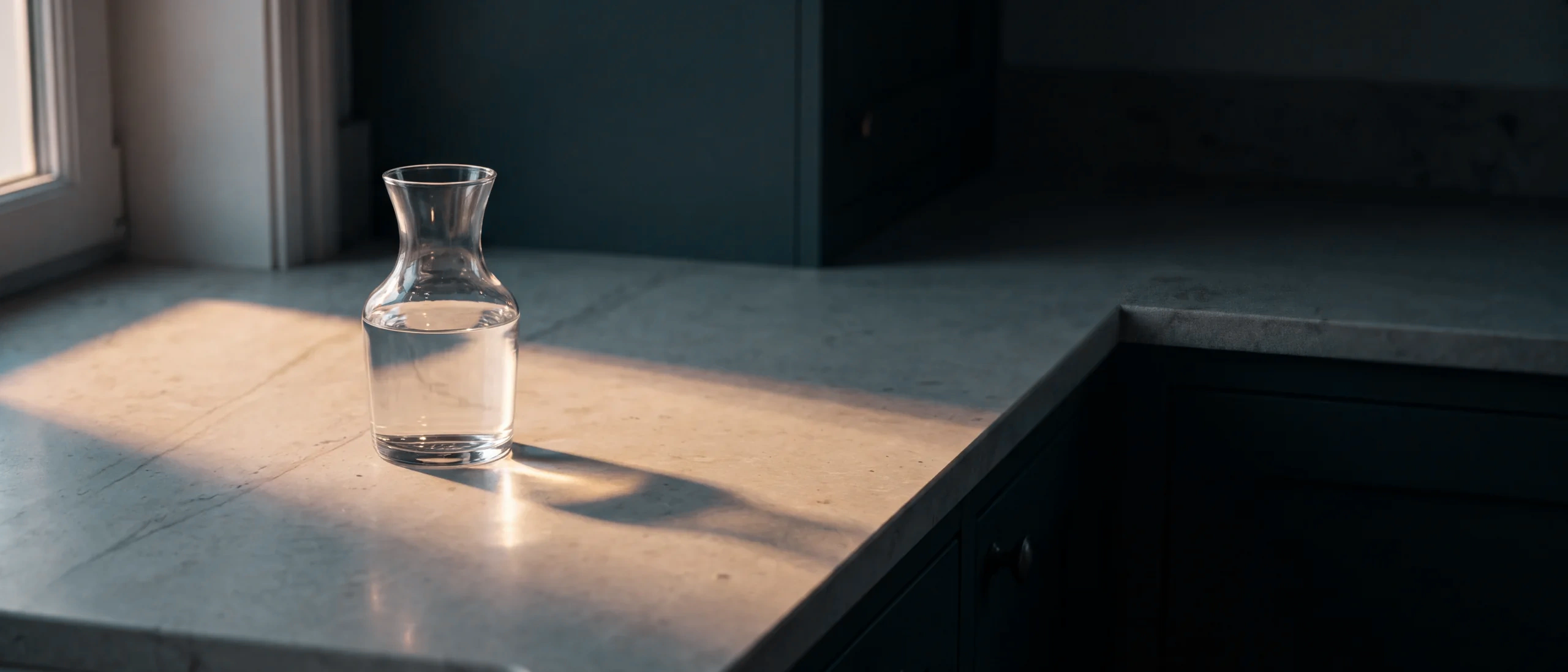 A glass carafe of water on a pale stone benchtop by a window, low warm sunlight casting its shadow across the stone, deep teal cabinetry behind.