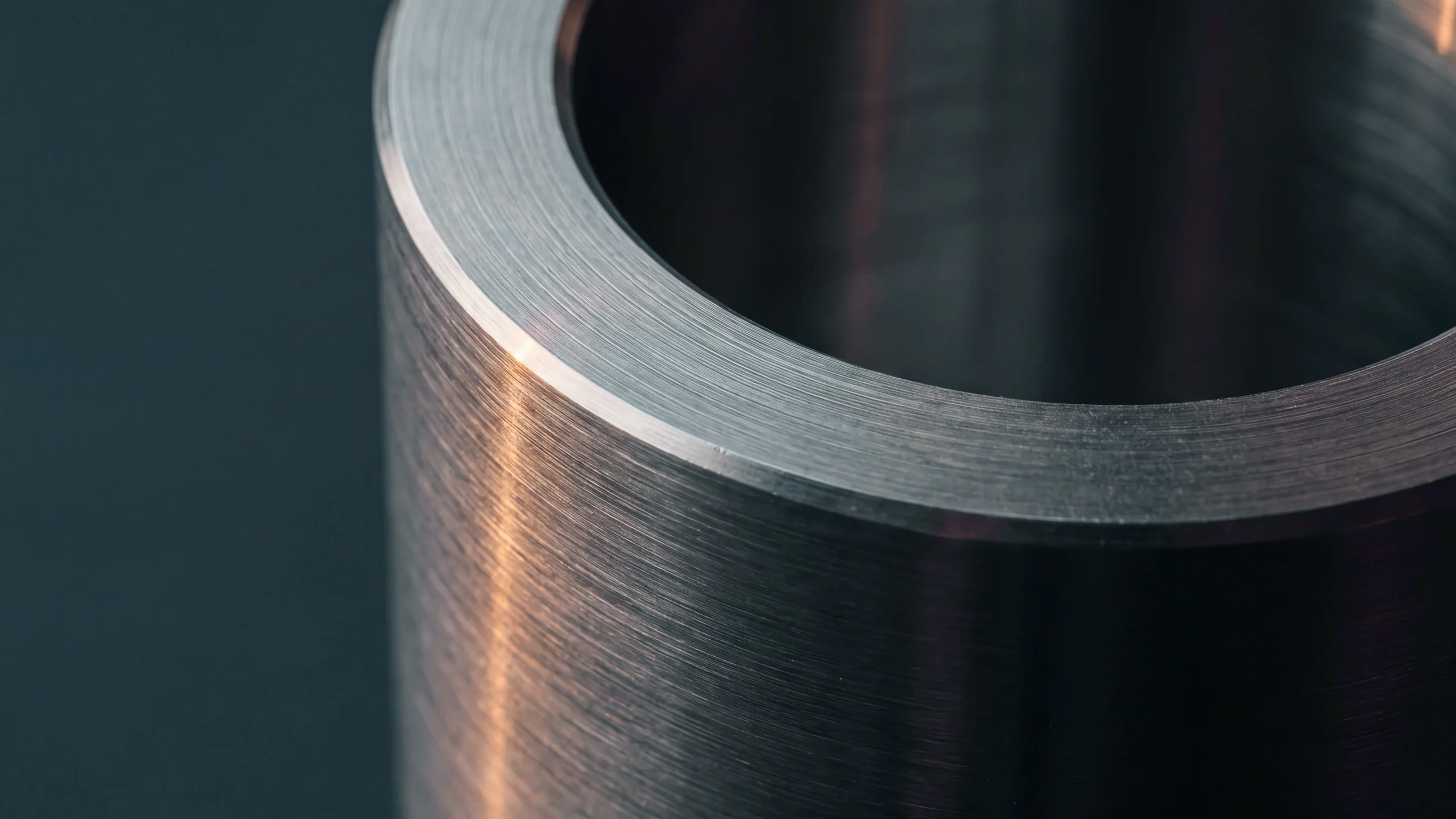 Brushed stainless steel in close-up, a machined rim and chamfer catching a low warm rake against a deep slate-teal ground.